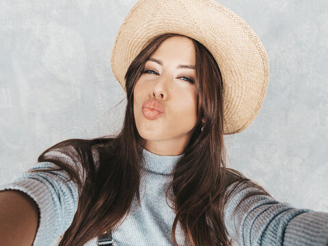 Portrait of cheerful young woman taking photo selfie and wearing modern clothes and hat. Beautiful holding smartphone camera. Smiling model posing near gray wall. Making duck face