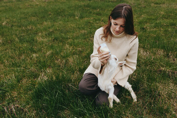 Naklejka premium A woman sits on grass in a quiet field, bottle feeding a white goat kid, sharing a gentle intimate moment with a small animal, capturing a warm lifestyle scene amid nature and open space