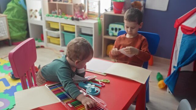 Two brothers reach for crayons and paper, hand on colored pencil set at a small red table in a playroom building; concentration.
