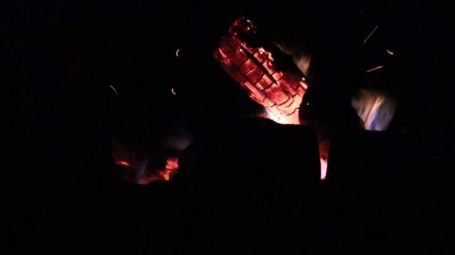 Close-up night shot of a bonfire with flames filling the frame. Burning wood, glowing embers, and flickering firelight visible against a dark background.