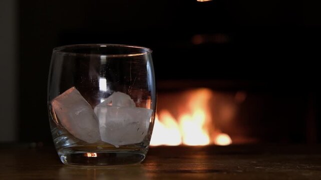 Close up ice and golden whiskey poured into glass in front of fireplace