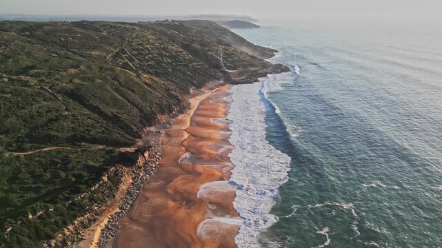 Slow motion aerial clip of Praia do Salgado in Nazare captures waves crashing against the rugged cliff and sandy beach during a bright day. Perfect for travel and nature projects
