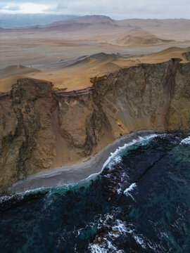 Aerial view of Reserva Nacional de Paracas with steep coastal cliffs, a narrow shoreline, and vast desert plains under a cloudy sky Paracas, Ica, Peru.