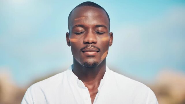Calm young African man with painted face meditating outdoors in white shirt with serene blue sky background