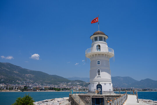 White lighthouse with Turkish flag in Alanya harbor on sunny day. Scenic Mediterranean port landscape, nautical navigation, historical landmark and summer travel concept in Turkey