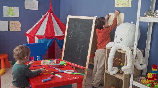 Boy drawing with crayons at a red table while brother pins artwork to an easel in a colorful playroom; creative concentration.