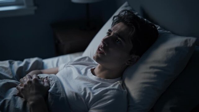Young man lying awake in bed at night looking worried and stressed with hands clasped under blanket