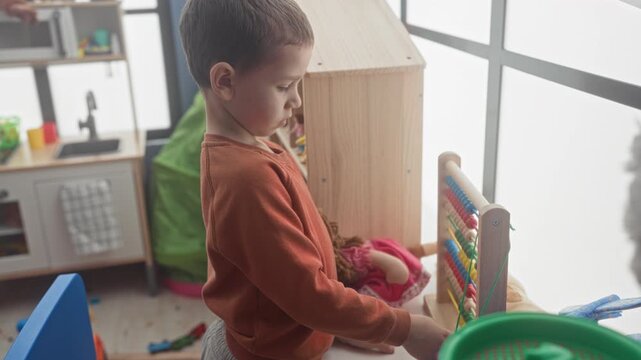 Boy playing with a wooden abacus, moving beads with his hand beside a doll in a bright playroom building; quiet concentration.