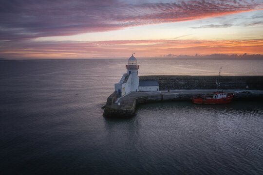 Aerial view of Balbriggan Lighthouse at the end of a stone pier with a red boat docked under a vibrant sunset sky Balbriggan, County Dublin, Ireland.