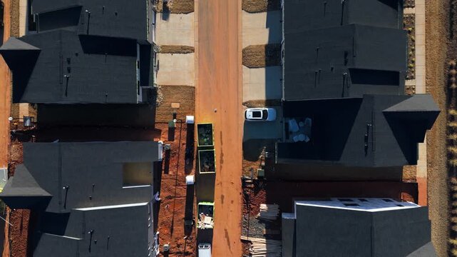 Overhead View Of Dark-roofed Townhouses Along Dirt Road With Waste Bins At Residential Construction Site. aerial topdown shot