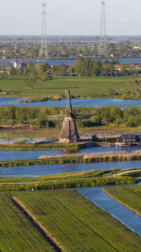 Aerial view of a historic Kinderdijk windmill standing amidst lush green fields and winding canals with power lines in the background Kinderdijk, South Holland, Netherlands.