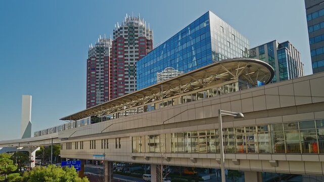 An elevated monorail station stands under a clear blue sky in a modern district. Tall residential towers and glass office buildings create a clean, urban cityscape in Tokyo.