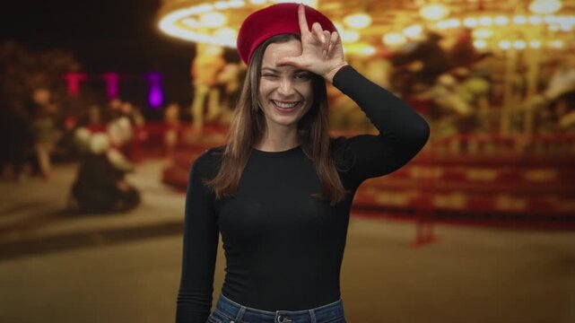 Woman in red beret and black longsleeve makes l sign on forehead, smiling broadly with tongue out in a night fair street with carousel lights behind her; playful mischief.