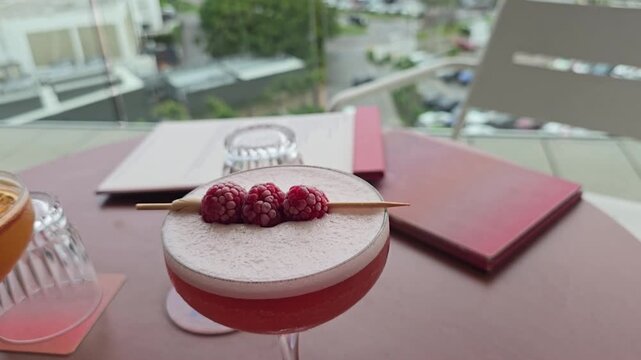 A cocktail sitting on a table with raspberries on a skewer placed on top, with menus and the city in the background at a roof top bar.