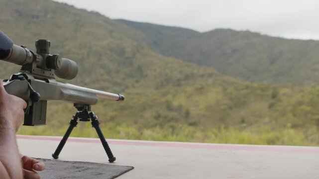 A sniper aims his rifle at a target below that is out of sight and pulls the trigger, a puff of smoke comes out of the barrel as the bullet is discharged with mountain range in background.
