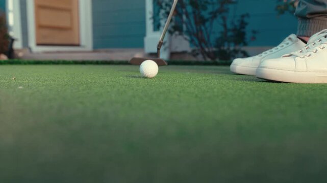 Slow motion of a putter hitting ball towards camera on outdoor backyard green and a residential house entry behind. Perfect for golf training videos, lifestyle content, and product ads.