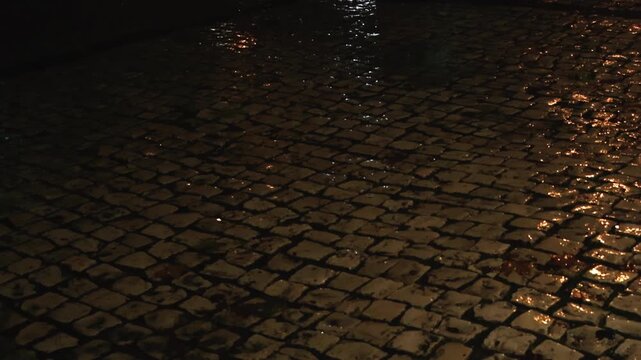 Heavy rain falling on the wet cobblestones of a traditional portuguese pavement at night, with city lights creating reflections and puddles on the dark, ancient street surface