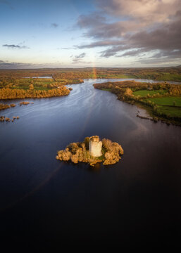 Aerial view of Cloughoughter Castle on a small island surrounded by calm lake waters and autumn foliage under a rainbow County Cavan, Ireland.