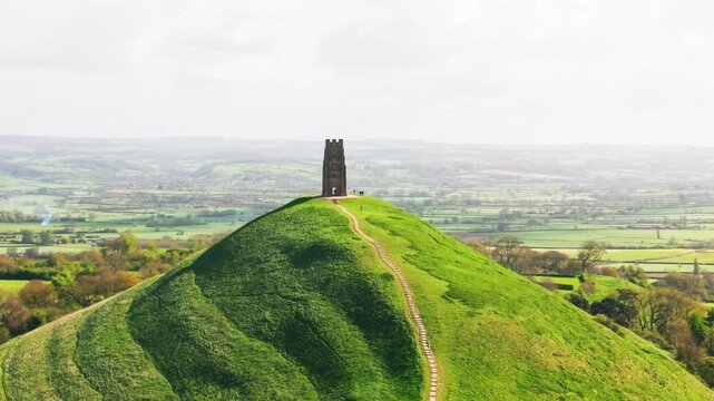 Drone view of Glastonbury Tor showing a winding footpath climbing a lush green hill towards St Michael&rsquo;s Tower with Somerset countryside stretching into the distance