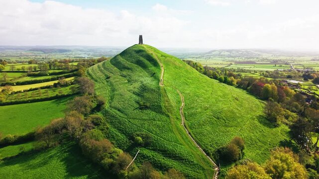 Aerial drone view approaching Glastonbury Tor showing the winding path up the green hill leading to the historic tower with surrounding Somerset countryside in the United Kingdom