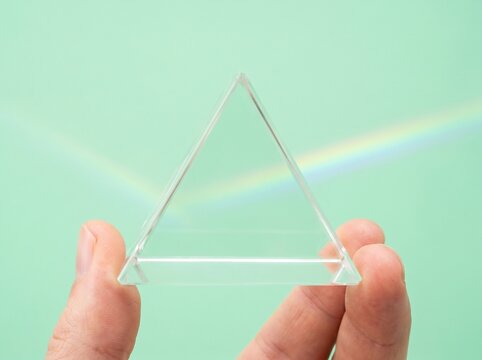 A hand holds a clear glass triangular prism, refracting light into a rainbow spectrum against a soft green background.