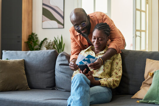 Black couple reviewing travel documents and checking smartphone while preparing for trip at home, planning vacation logistics. Useful for travel booking, tourism marketing, trip planning