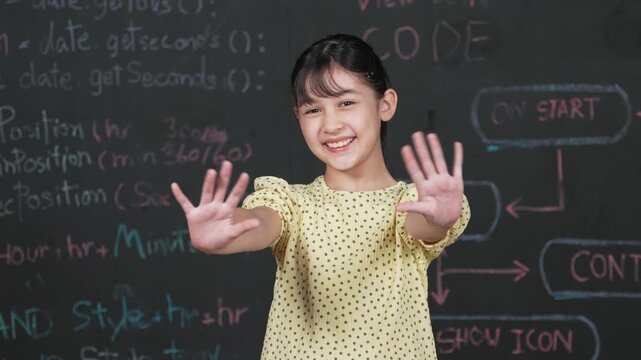 Cute happy girl waving to camera while standing at blackboard with engineering prompt or coding, programming system written in STEM technology classroom. Smiling student looking at camera. Edification