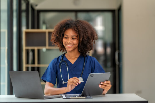 Smiling African American female doctor using digital tablet and laptop at clinic desk.