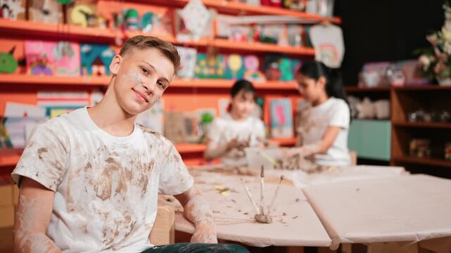 high school boy looking at camera while multicultural children modeling clay. Diverse student having pottery class together. Child smile while wearing white shirt with mud stained cloth. Edification
