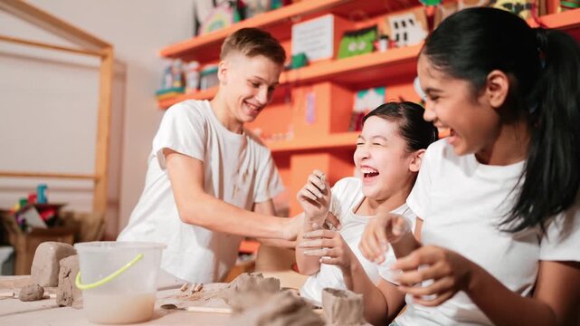 Handsome student playing with friend while put the clay on shirt at class. Group of diverse children working or modeling vase. Happy boy put water to young girl while laugh with happy. Edification