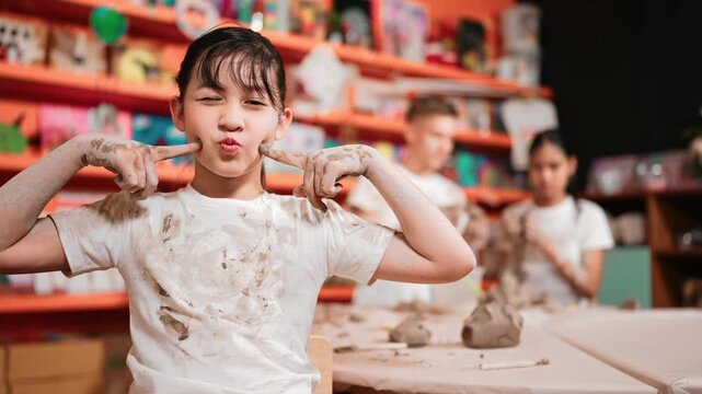 Asian girl pose at camera while diverse children modeling clay behind. Happy cute student wearing dirty shirt while looking at camera at workshop in art lesson. Blurring background. Edification