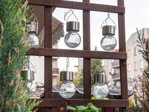 Set of glass solar garden lanterns hanging on wooden trellis near residential buildings, showing outdoor decoration, backyard lighting and eco friendly design detail.