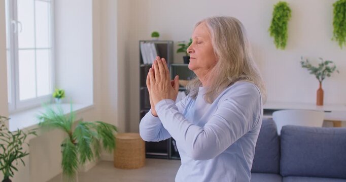 Mature grey haired woman meditating at home in living room, keeps hands in zen gesture, finding balance, breathing deeply, trying to relax, practicing yoga. Old people and meditation concept