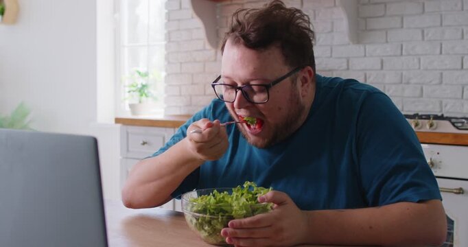 Plus size man eating salad to lose weight, person with obesity sitting with laptop at kitchen table to eat healthy food with with appetite. Young overweight person in glasses enjoying fresh vegetables