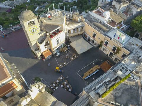 Aerial view of the historic Piazzetta square with its yellow clock tower, people walking and white cafe umbrellas in Capri, Campania, Italy.