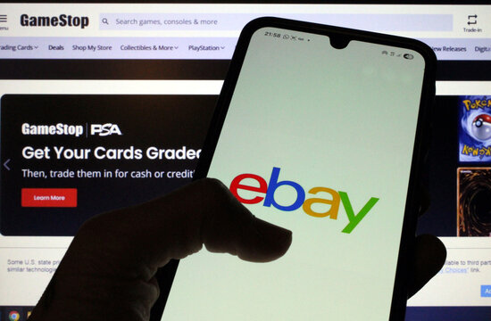 Hand holding smartphone with eBay logo in front of GameStop website, symbolizing online commerce, digital markets, and potential corporate acquisition trends
