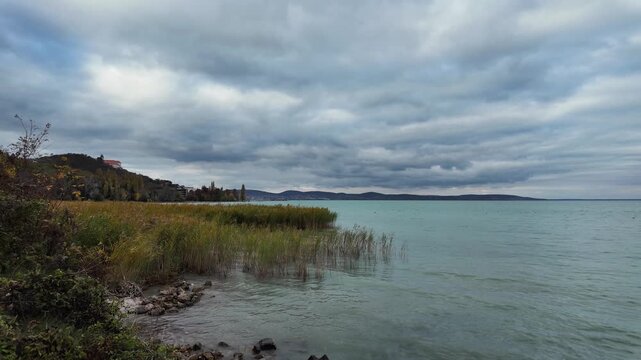 The famous Benedictine monastery on the Tihany peninsula on a cloudy autumn afternoon. Wavy Lake Balaton water with reeds and rocks in the foreground. Calm scenic slow motion landscape in Hungary.