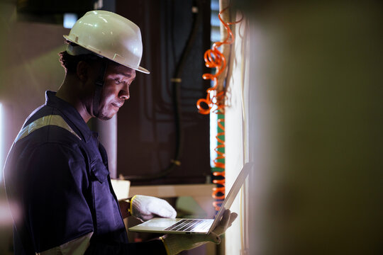 Black engineer using laptop to check machinery in factory