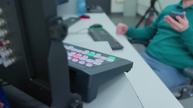 An engineer stops talking on a cell phone and go for a job in a TV control center. He sits at a keyboard and control panel on a desk