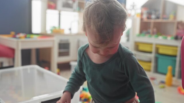 Little boy leans over clear plastic bin, picks up colorful toy with one hand and drool on chin in a building; play exploration curiosity.