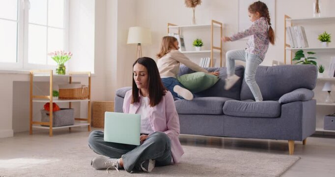 Mother sits on floor working with laptop, little kids play around making noise, home career mom suffering from mess working on computer, nursing children, freelancing, distracted self-employed