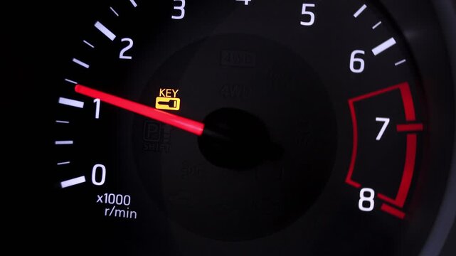 Extreme close-up of a vehicle tachometer needle moving up and down as the engine revs. The dashboard features glowing white RPM numbers and a yellow key warning indicator light.