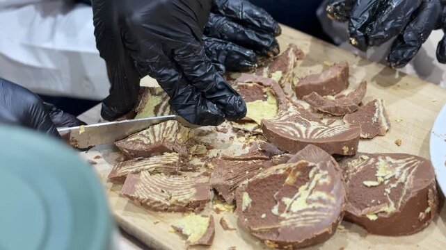 a person wearing black gloves slicing a chocolate biscuit mosaic cake on a wooden board. Professional food preparation showing the internal texture of the sweet dessert.
