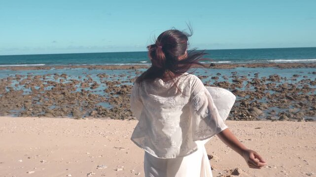 Handheld shot of a young Indonesian woman walking toward the sea on a rocky beach, then turning around and looking at the camera with a smile.
