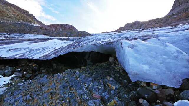 A dynamic low-angle shot showing a receding glacier, with a visible ice cave opening and a mix of blue ice and rugged moraine rocks in the foreground. The scene highlights the effects of climate chang