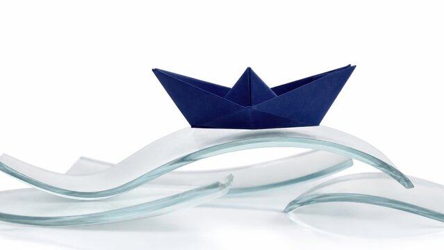 Modern conceptual photography of a small dark blue paper boat sitting on curved pieces of smooth frosted clear glass acting as abstract water waves