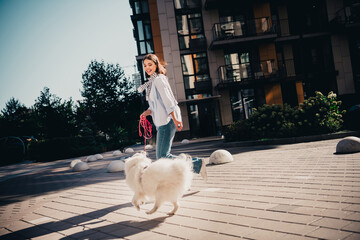 Young woman walking a fluffy white dog on a sunny city sidewalk near modern apartment buildings, smiling and enjoying an active urban stroll © deagreez