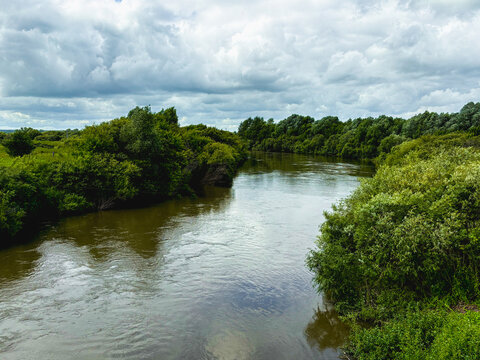 River. A picturesque summer landscape with a wide river, dense green trees on the banks under a cloudy sky.