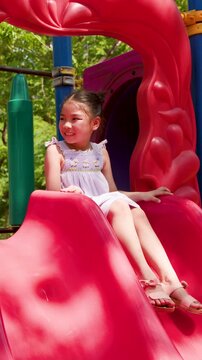 Happy Young Asian Girl Sliding Down Red Slide at Outdoor Playground