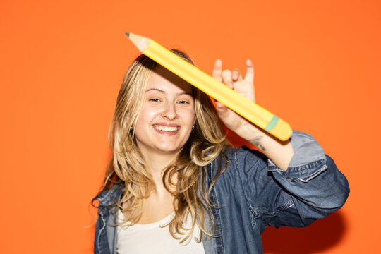 Smiling student holds giant pencil on orange background